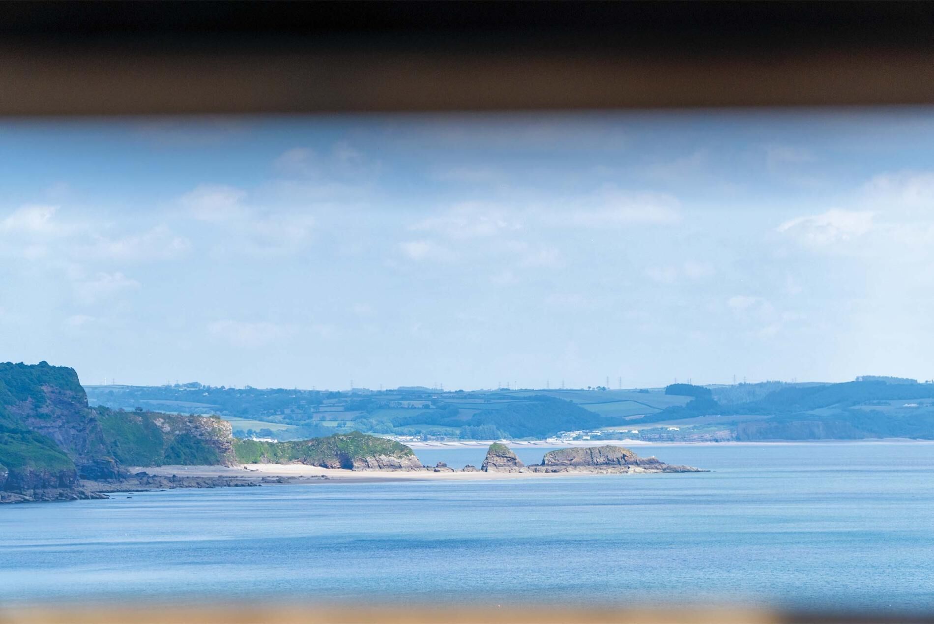 Eine Küstenlandschaft mit einer markanten Felsformation im Hintergrund, einem Sandstrand und sanften Hügeln.