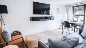 A modern living room with a flat-screen TV mounted on the wall, a dining area with a table and chairs, and a large window with curtains. - The Cottage - 2 Bedroom Cottage - Tenby (Tenby)
