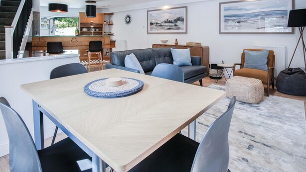 A modern living room with a dining area, featuring a wooden table, grey sofas, and a staircase leading to an upper level. - The Cottage - 2 Bedroom Cottage - Tenby (Tenby)