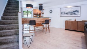 A modern kitchen with a glass island, wooden chairs, and a dining area with a wooden cabinet and a painting on the wall. - The Cottage - 2 Bedroom Cottage - Tenby (Tenby)