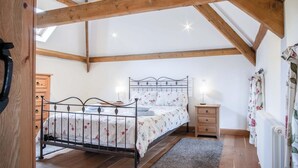 Une chambre avec un plafond en pente, des poutres en bois, un cadre de lit en métal, une table de chevet et une commode, ainsi qu’un radiateur.