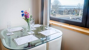 A dining table with a glass top, set with white napkins, silverware, and wine glasses, positioned by a window with a view of buildings and a tree. - Harbour Hideaway - 1 Bedroom Apartment - Saundersfoot (Saundersfoot)