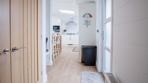 En smal gang fører til et køkken med hvide skabe, en mikroovn og et spiseområde med stole.