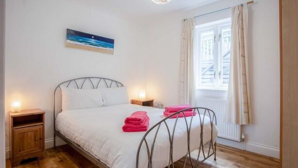 A bedroom with a metal bed frame, white bedding, two bedside tables with lamps, a painting of a beach scene, and a window with curtains. - Kensington Apartment - 2 Bedroom - Saundersfoot (Saundersfoot)
