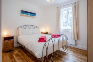 A bedroom with a metal bed frame, white bedding, two bedside tables with lamps, a painting of a beach scene, and a window with curtains. - Kensington Apartment - 2 Bedroom - Saundersfoot (Saundersfoot)