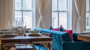 A dining area with a wooden table set for a meal, a blue sofa with colorful pillows, and large windows with curtains. - Kensington Apartment - 2 Bedroom - Saundersfoot (Saundersfoot)