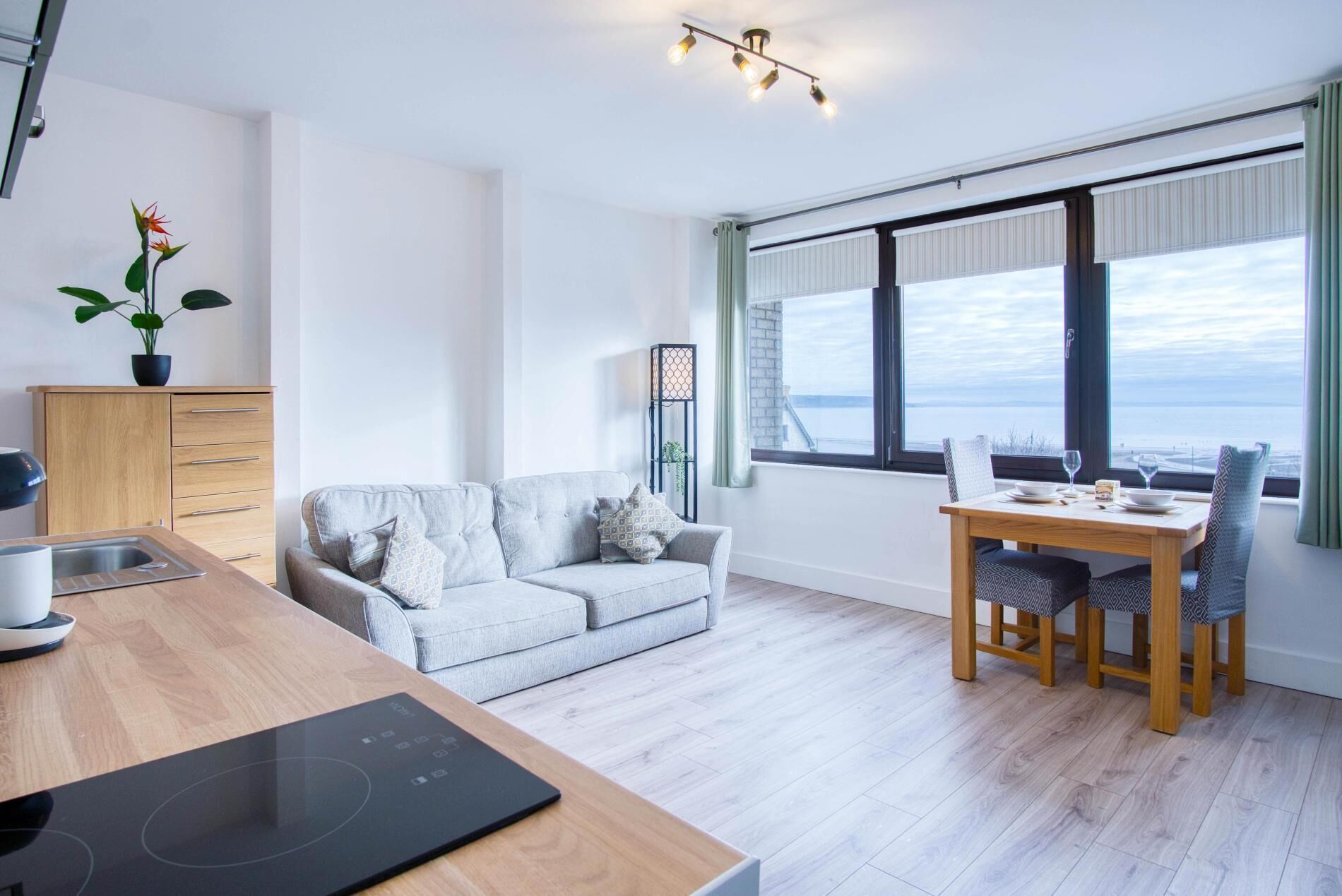 A modern living room with a grey sofa, a wooden dining table with chairs, a built-in kitchen with a sink, and a large window with a view of the sea.