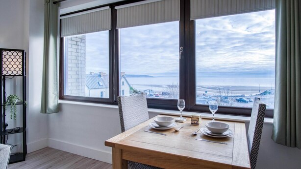 A dining area with a wooden table and chairs, a window with a view of the sea and buildings, and a small plant on a shelf.