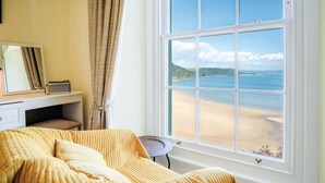 A bedroom with a bed, a desk, a chair, and a large window overlooking a beach. - 5 Belle Vue Court - 2 Bedroom Apartment - Tenby (Tenby)