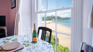 A room with a large window overlooking a beach, a table with a bottle of champagne, and a chair. - 5 Belle Vue Court - 2 Bedroom Apartment - Tenby (Tenby)