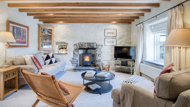 A cozy living room with a stone fireplace, wooden beams, a sofa, a wicker chair, and a TV. - Fern Cottage - 2 Bedroom Cottage - St Florence (Tenby)