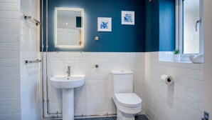 A bathroom with white tiles, a blue accent wall, a sink, a toilet, and framed artwork. - The Studio - 1 Bedroom Cottage - Princes Gate (Narberth)