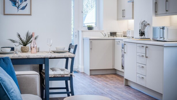 A modern kitchen with white cabinetry, a dining area with a wooden table and chairs, and a view of the outdoors through a window. - The Studio - 1 Bedroom Cottage - Princes Gate (Narberth)