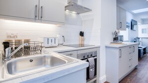 A modern kitchen with white cabinetry, a stainless steel sink, a built-in oven, and a compact dining area with a table and chairs. - The Studio - 1 Bedroom Cottage - Princes Gate (Narberth)