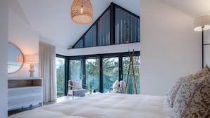 A modern bedroom with a large bed, a desk, and a chair. There is a large window with a view of trees.