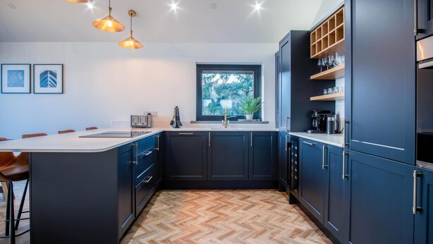 A modern kitchen with dark cabinetry, a herringbone wood floor, pendant lighting, and a window with a view of trees.