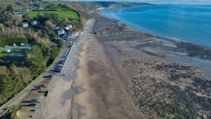 An aerial view of a coastal area with a sandy beach, a road with parked cars, and houses along the coastline.
