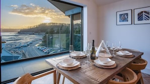 A dining table set with plates, glasses, and a bottle, overlooking a coastal road with cars and trees.
