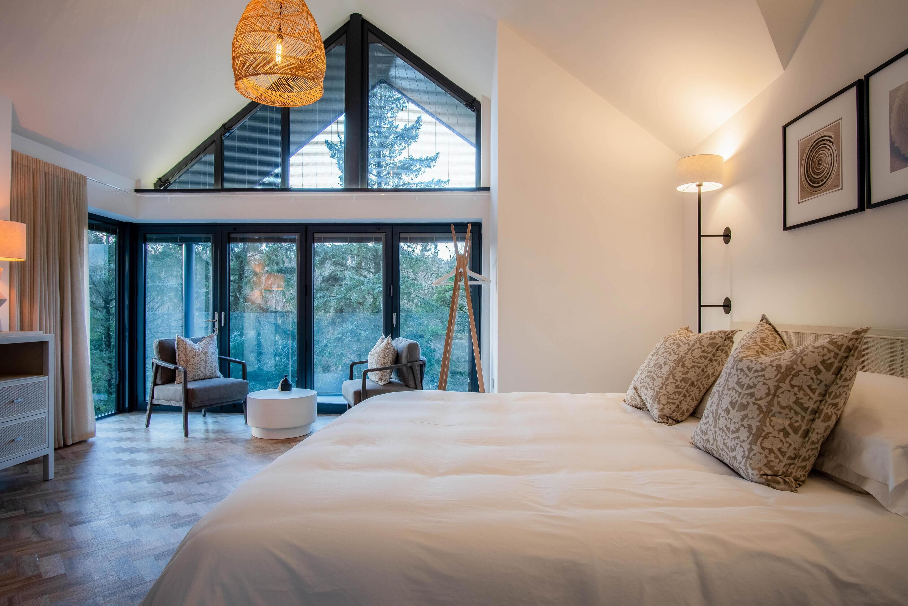 A modern bedroom with a large bed, two chairs, a side table, and a large window with a view of trees.