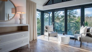 A modern living room with a herringbone wood floor, a white console table, a round mirror, and a large window offering a view of trees.