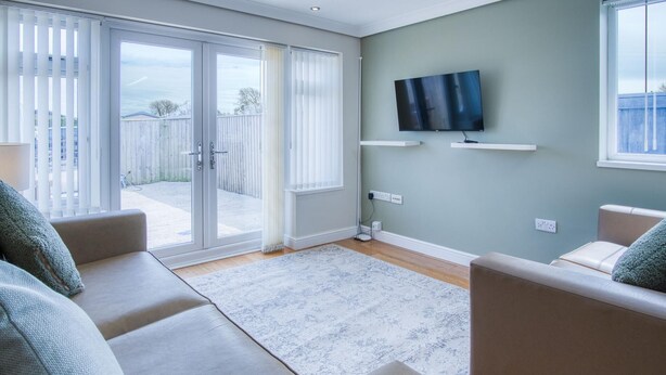 A living room with a sofa set, a flat-screen TV mounted on the wall, and a sliding glass door leading to an outdoor area. - Ridgeway Hideaway - 1 Bedroom Holiday Home - Manorbier (Tenby)