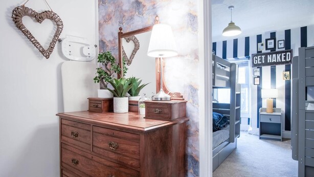 Een kamer met een houten dressoir, een spiegel en een lamp. In de achtergrond staat een bed met een bord waarop staat: GET NAKED.