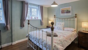 A bedroom with a white metal bed frame, floral bedding, a bedside table with a lamp, and a window with curtains. - Beachwood - 2 Bedroom Cottage - Penally (Tenby)