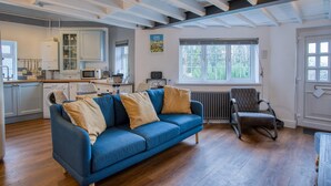 A modern kitchen with white cabinets, a microwave, and a washing machine. There is a blue sofa with cushions, a radiator, and a window with a view of greenery. - Beachwood - 2 Bedroom Cottage - Penally (Tenby)