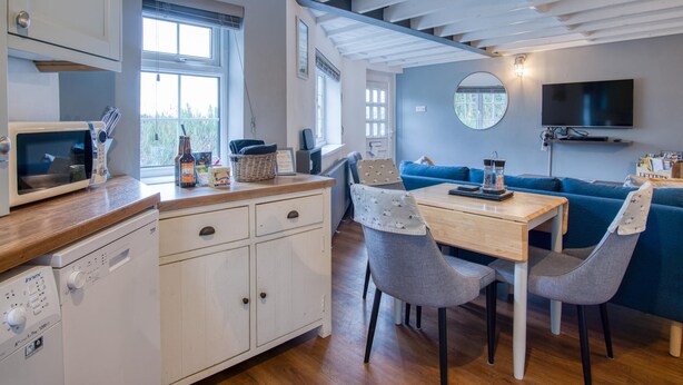 A compact kitchen with a microwave, a coffee machine, and a sink. There is a dining area with a table and chairs, and a blue sofa in the background. - Beachwood - 2 Bedroom Cottage - Penally (Tenby)