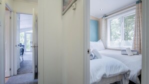 A hotel room with two beds, a window with blinds, a door leading to a hallway, and a painting on the wall. - 1 Manor Lodge - 3 Bedroom Lodge - Pendine (Carmarthen)