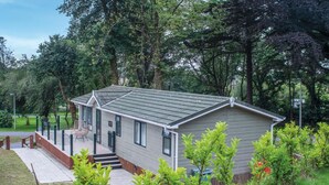 A small, single-story mobile home with a grey roof and white siding, surrounded by lush greenery and trees. - 1 Manor Lodge - 3 Bedroom Lodge - Pendine (Carmarthen)