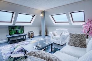 Een moderne woonkamer met een flatscreen-tv, een salontafel, een bank en een fauteuil. Er zijn dakramen en een groot raam dat zorgt voor natuurlijk licht.