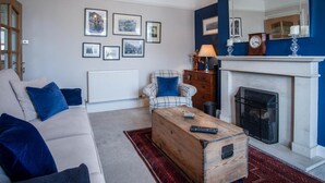A living room with a sofa, armchair, wooden chest, fireplace, and framed pictures on the wall.