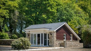 A small, single-story building with a sloped roof, stone foundation, and a covered porch with glass doors. - The Pig Sty - 1 Bedroom Studio - Princes Gate (Narberth)