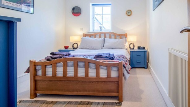 A bedroom with a wooden bed, two bedside tables with lamps, a window, and a radiator. - 29 Rhodewood House - 2 Bedroom - Saundersfoot (Saundersfoot)