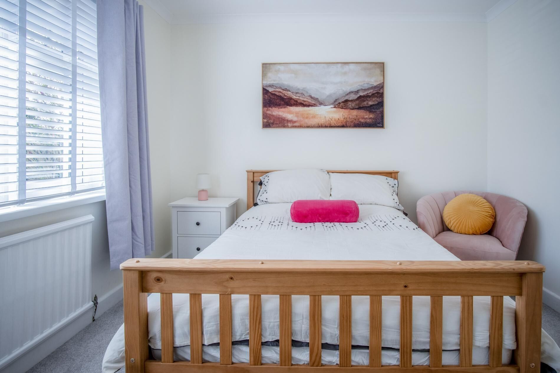 A bedroom with a wooden bed, white bedding, a nightstand with a lamp, a chair with a cushion, and a painting on the wall.