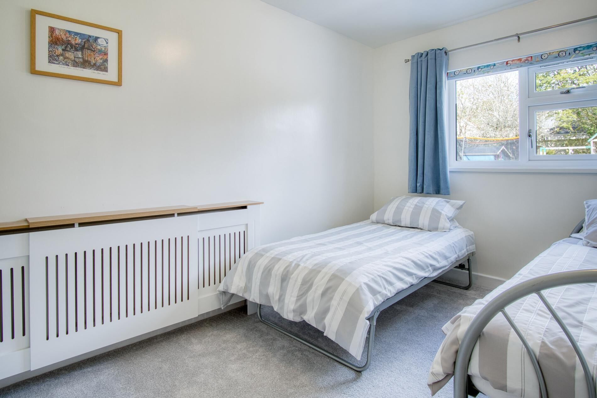 A bedroom with a single bed, a radiator, a window with curtains, and a framed picture on the wall.