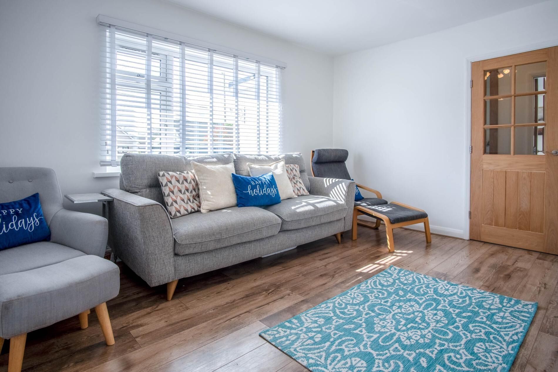 A modern living room with a grey sofa, armchairs, and a wooden bench. There is a wooden door and a window with blinds.