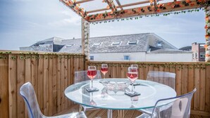 A glass-top table with three wine glasses, surrounded by clear plastic chairs, set against a wooden fence with decorative foliage.