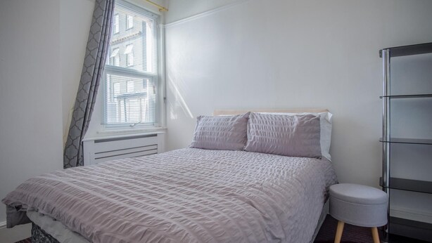 A bedroom with a bed, a bedside table, a chair, a radiator, and a window with curtains.