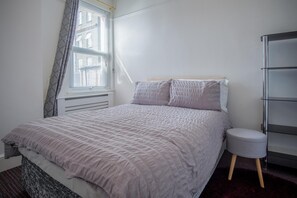 A bedroom with a bed, a bedside table, a chair, a radiator, and a window with curtains. - Skyfall - 2 Bedroom Apartment - Milford Haven (Milford Haven)
