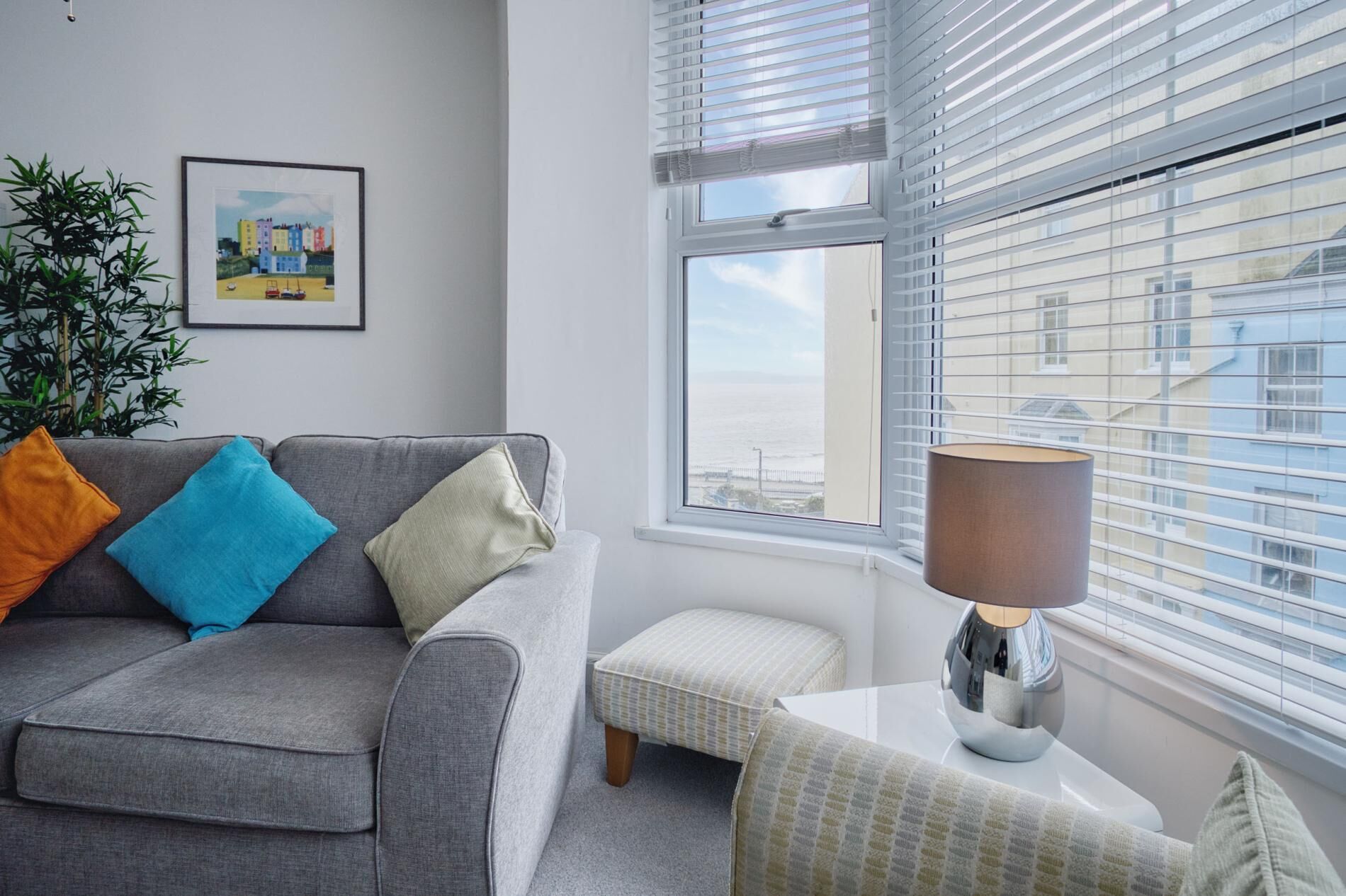 Een moderne woonkamer met een grijze bank, een klein voetenbankje, een lamp en een lijst met een afbeelding aan de muur.