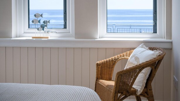 Une chambre à coucher avec un lit, un fauteuil en osier doté d’un coussin, et une vue sur l’océan par deux fenêtres.