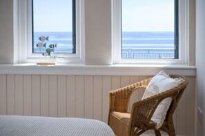 Een slaapkamer met een bed, een rieten stoel met kussen en uitzicht op de oceaan door twee ramen.