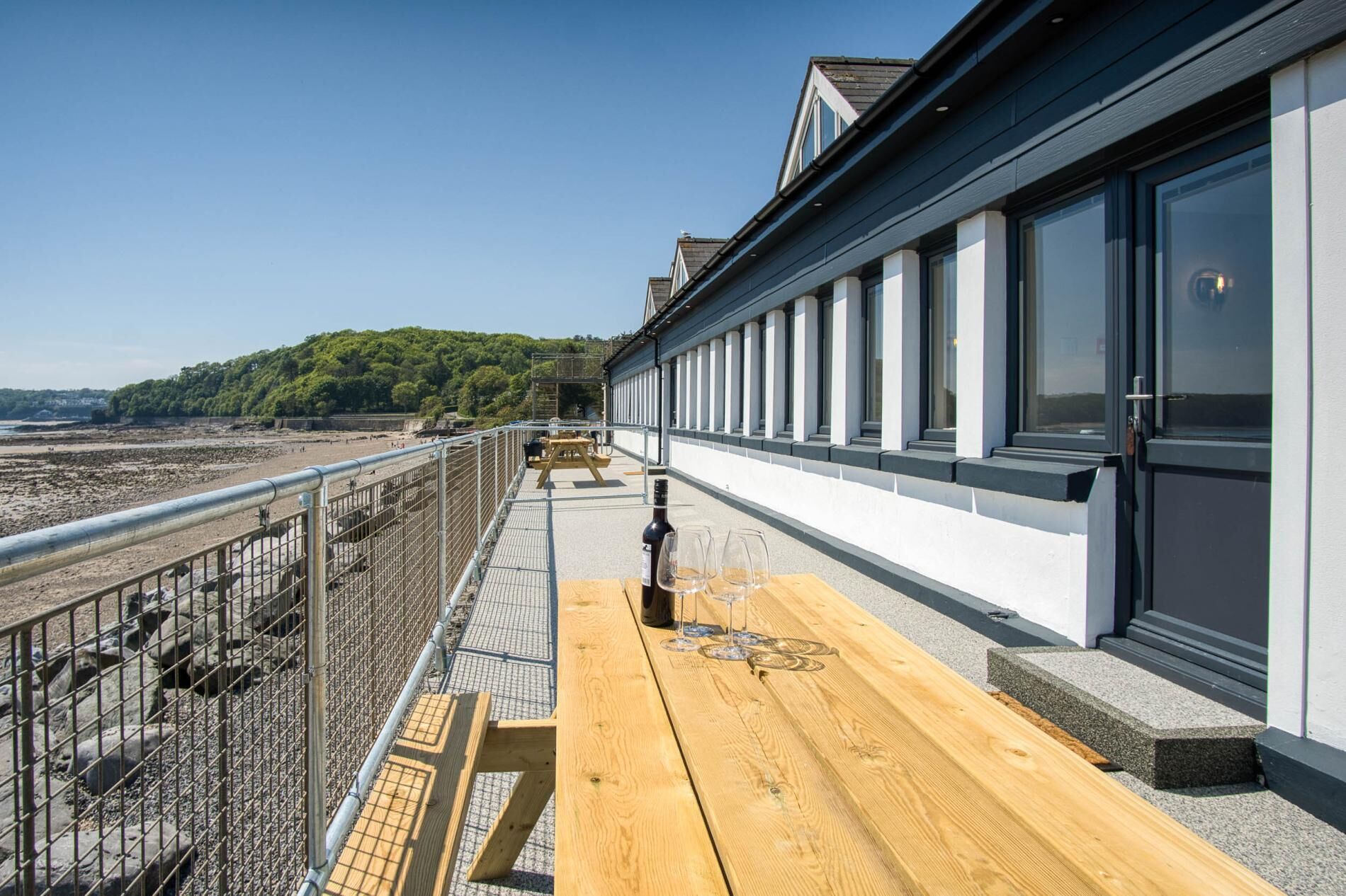 A wooden deck with a bottle and glasses, overlooking a beach and a building with large windows.