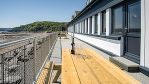 Une terrasse en bois avec une bouteille et des verres, donnant sur une plage et un bâtiment doté de grandes fenêtres.