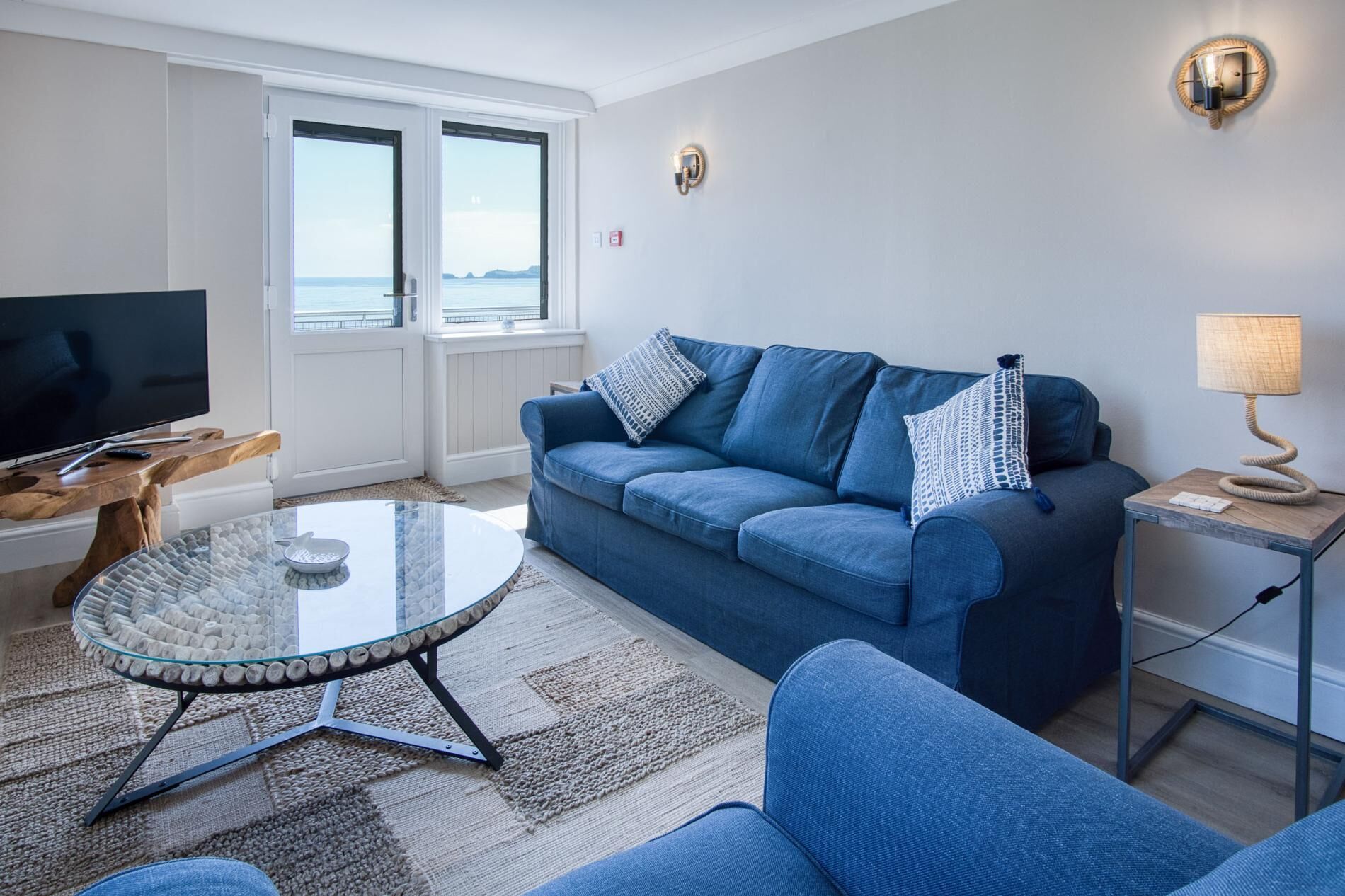 A living room with a blue sofa, a glass coffee table, a TV on a wooden stand, and a lamp on a side table.