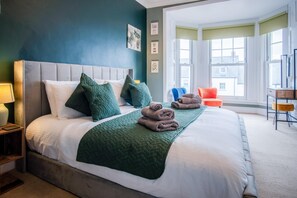 A neatly made bed with a green quilt, pillows, and folded towels. There are framed pictures on the wall, a window with a view, and a chair by the window. - The Lanterns - 9 Bedroom - Sleeps 17 - Saundersfoot Village Centre (Saundersfoot)
