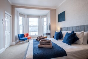 A hotel room with a large bed, a blue and white bedspread, multiple pillows, a chair, a desk, and a large window with a view of buildings. - The Lanterns - 9 Bedroom - Sleeps 17 - Saundersfoot Village Centre (Saundersfoot)