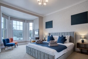 A modern bedroom with a large bed, a seating area with chairs, and a view through large windows. - The Lanterns - 9 Bedroom - Sleeps 17 - Saundersfoot Village Centre (Saundersfoot)